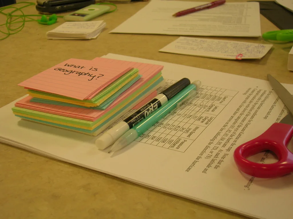 Une pile de fiches bristol de couleur posée sur une feuille imprimée, la première porte au feutre la question « what is geography? », à côté de deux stylos et d’une paire de ciseaux rouges.