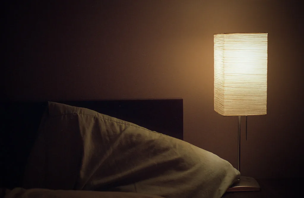 Une lampe de chevet en papier encore allumée la nuit, à côté d’un lit vide et défait.