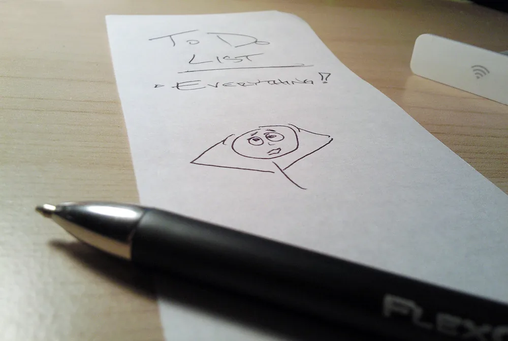 Une feuille posée sur un bureau, titrée « To Do List », avec une seule ligne : « Everything! », et un petit bonhomme dessiné à l’air dépassé. Un stylo est posé devant.
