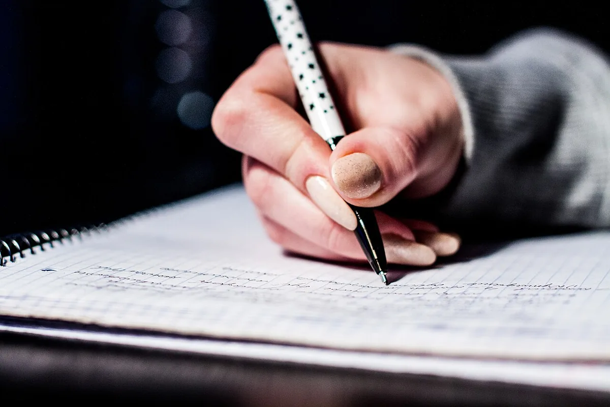 Close-up of a hand writing in a spiral notebook on squared paper, under a soft desk light against a dark background.