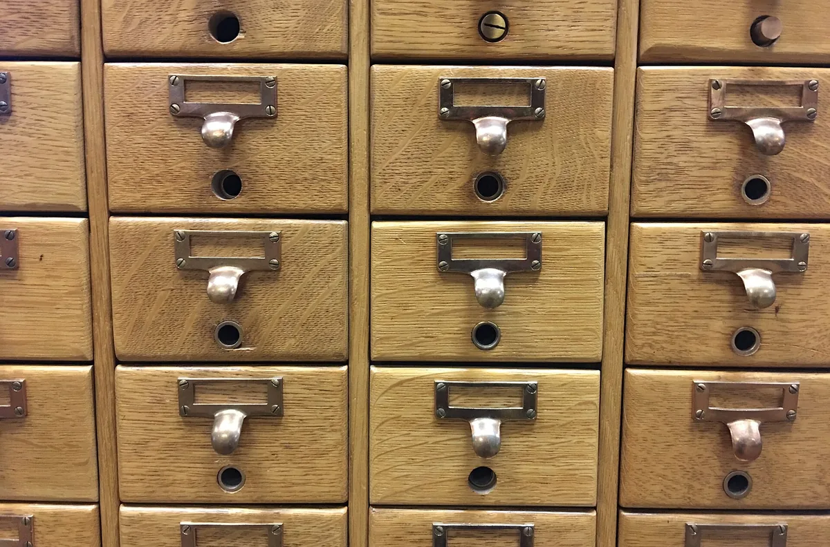Un mur de tiroirs à fiches en bois, chacun portant une étiquette manuscrite dans un porte-étiquette métallique.