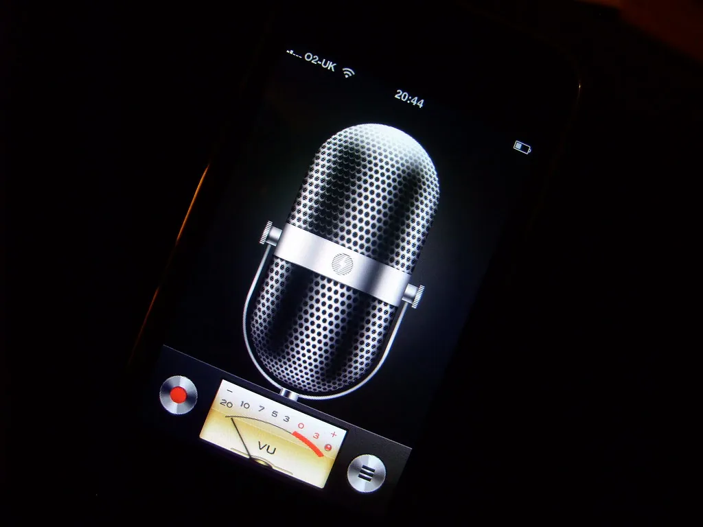 Un des premiers iPhone affichant l’ancienne app de mémos vocaux, un grand micro chromé sur fond noir.