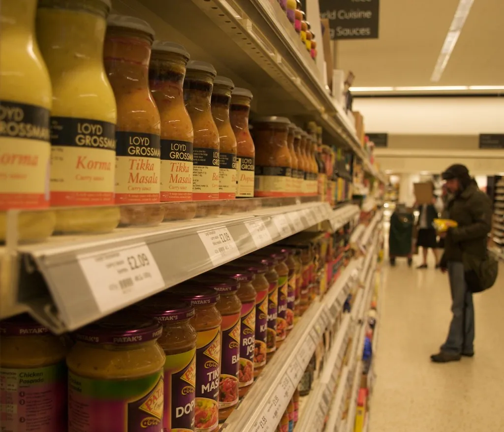 Gros plan sur une étagère de supermarché remplie de rangées de bocaux de sauce au curry, korma, tikka masala, avec leurs étiquettes de prix, et un client debout plus loin dans l’allée.